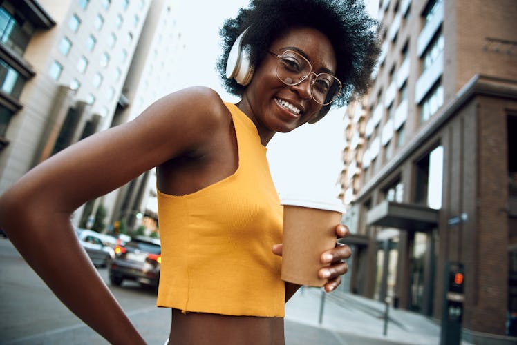 Waist up portrait of smiling woman with headphones holding coffee while walking on the street and li...