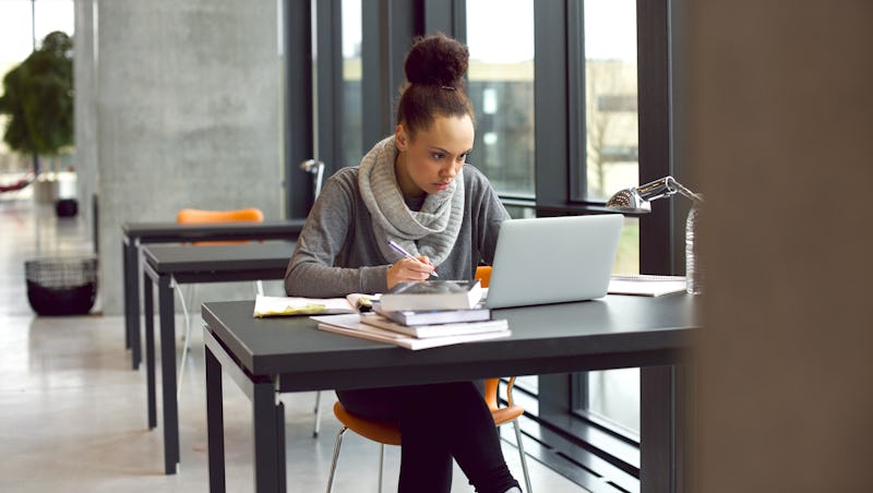 Young afro american woman sitting at table with books and laptop for finding information. Young student taking notes from laptop and books for her study in library.
