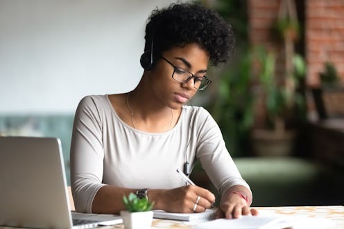 Serious african female smart student sitting at table wearing headphones listening online lecture wr...