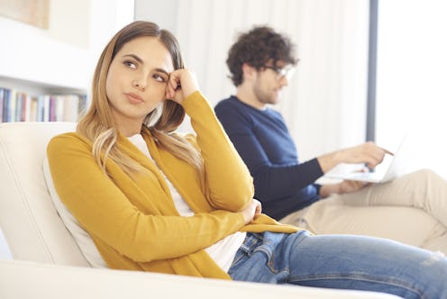 Shot of a careworn young woman sitting in couch while young man using laptop in the background.