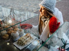 Smiling blond woman is curious looking at the apple candies on the other side of the window, she is ...