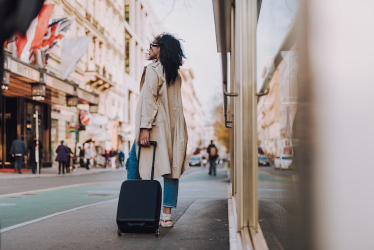 Full length portrait of beautiful young lady in elegant trench coat holding handle of black travel s...
