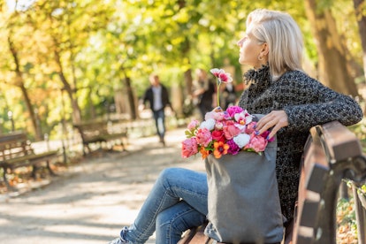 Portrait of young blonde dreamy woman with bouquet of flowers in backpack sits on the bench, side vi...