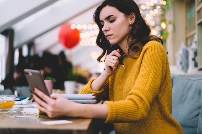 Side view of thoughtful young woman with brown hair and smartphone in yellow sweater touching chin while sitting at desk with notebook in cafe