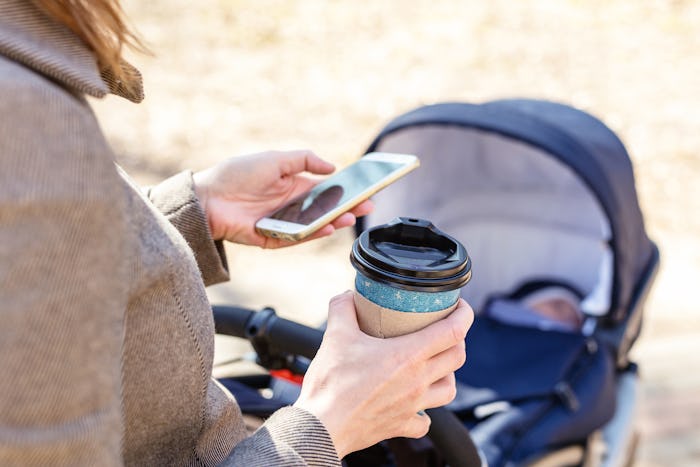 Woman holding cup of coffee to go and using smartphone during walk with baby carriage. Modern young ...