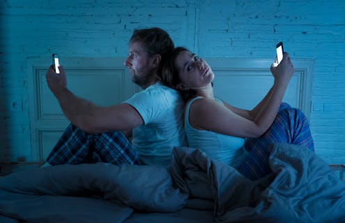 Sad man and woman married couple using their smart mobile phone in bed at night ignoring each other ...