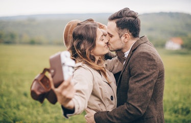 A happy couple kisses and hugs while taking a selfie with a camera in the countryside.