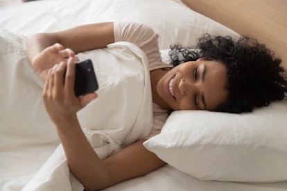 Close up african woman lying in comfortable bed covered with white blanket holding mobile phone chat...