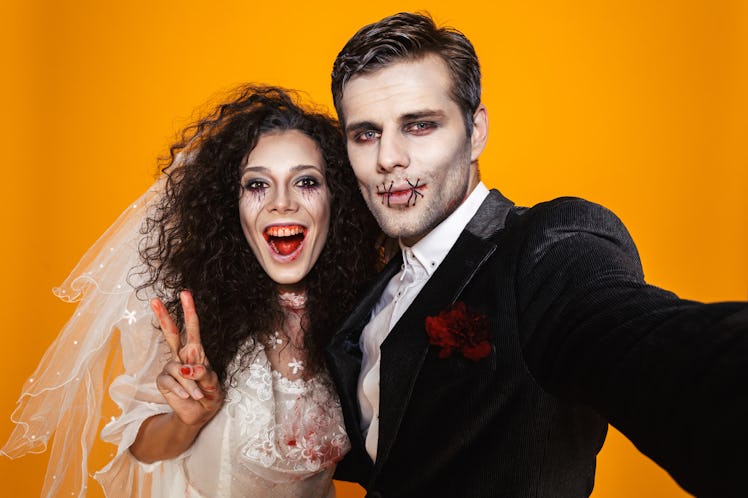Funny pretty zombie bride and groom in halloween costumes making selfie at camera and smiling isolat...