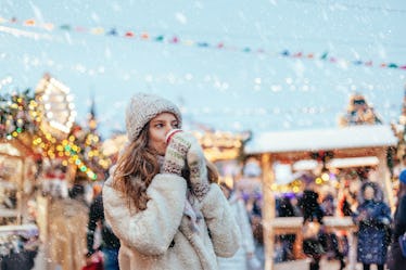 A woman dressed in a cap, fluffy coat, and gloves drinks hot chocolate at snowy holiday market.