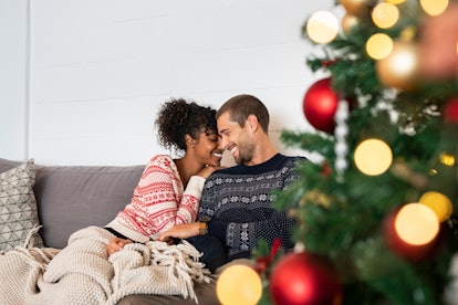 A happy couple relaxes on a comfortable couch wearing holiday sweaters near a Christmas tree.