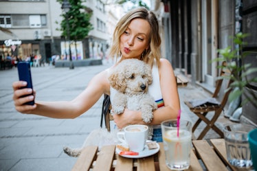 A blonde woman takes a picture with her dog while eating at a dog-friendly brunch spot in NYC.