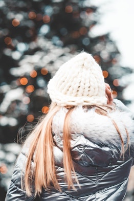 A blonde woman faces a holiday tree at the market in a metallic coat and knit hat.