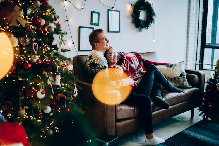 A guy in a festive sweater cuddles with his girlfriend on the couch, watching a romantic holiday mov...