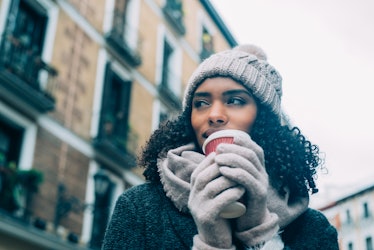 A woman in the city holds a cup of hot chocolate and wears chic winter clothes.