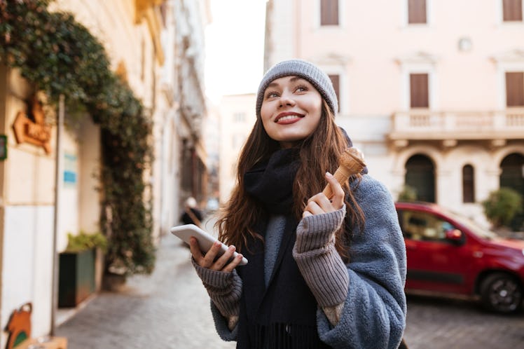 Cheerful cute young woman using cell phone and eating ice cream in the city