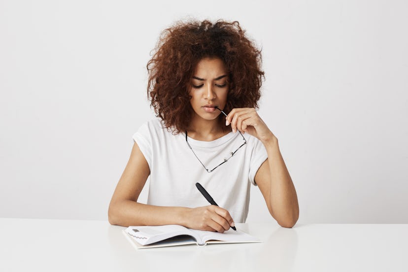 African girl thinking writing in notebook sitting at table over white background. Copy space.