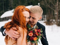 A bride with red hair holds a bouquet of red flowers while laughing with her groom during a holiday ...