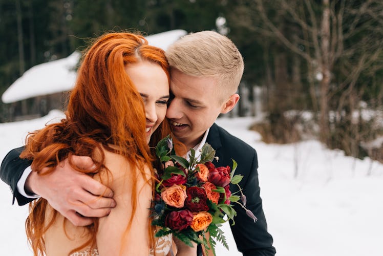A bride with red hair holds a bouquet of red flowers while laughing with her groom during a holiday ...
