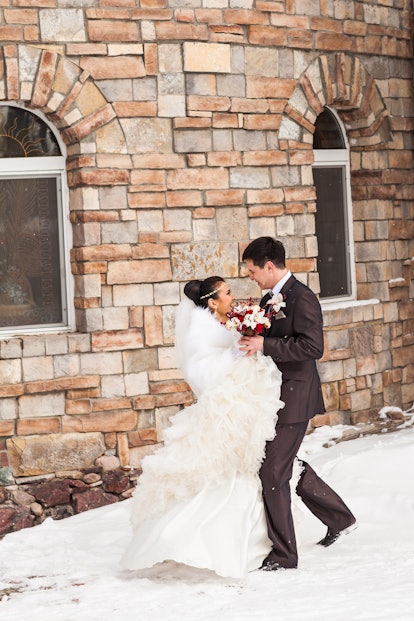 A happy, newly married couple laughs in the snow next to a brick wall on their wedding day.