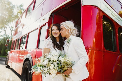 A couple stands next to a red double-decker bus with a bouquet of flowers during their holiday weddi...