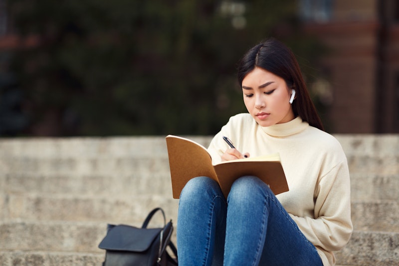 Girl in airpods writing in diary, preparing for lecture, sitting on university stairs in campus