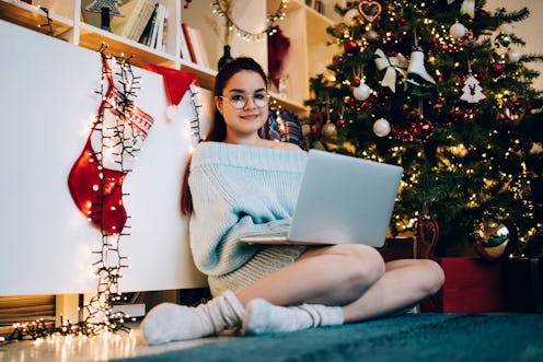 Pleased modern woman in glasses and warm sweater typing on laptop while sitting beside decorated Chr...