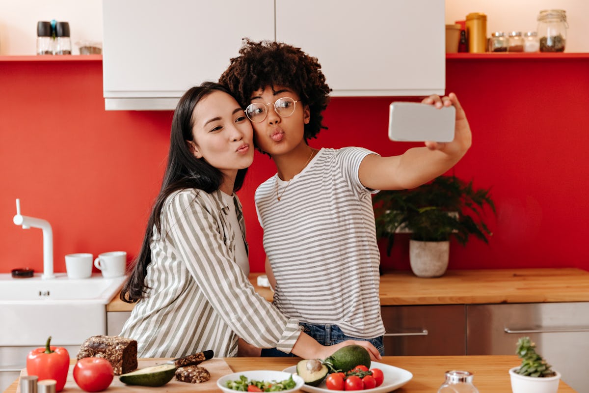 Brown-eyed girls blows kiss against background of kitchen. Women take selfie while cooking salad
