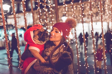 A woman and her French bulldog stand under twinkly lights at an outdoor holiday market.