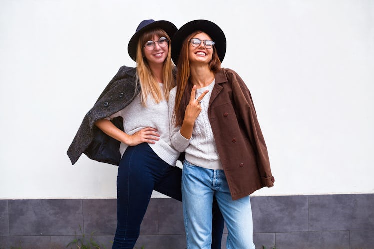 Two happy women pose in front of a white wall, wearing jeans, felt hats, fall jackets, and knit swea...