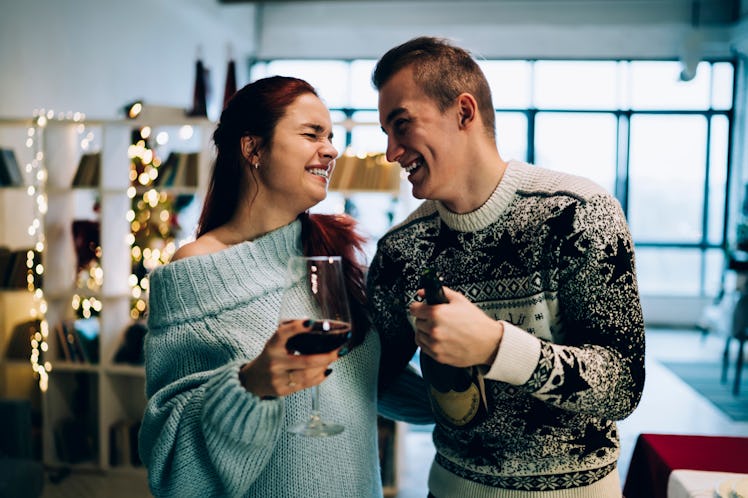 A happy couple wearing cozy sweaters and holding wine laughs while hugging each other in a cozy apar...