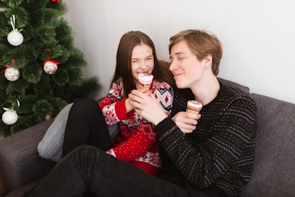 A happy couple wearing holiday sweaters sits on a sofa at home while eating ice cream next to a Chri...