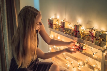A blonde woman sets up little holiday gift boxes and string lights on shelves in her home.