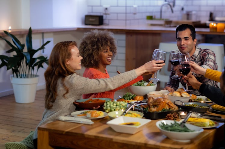 A group of friends in a kitchen laugh as they toast their wine glasses over the table of Thanksgivin...