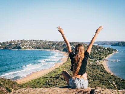 A girl sits on mountain overlooking the ocean with her arms up in Sydney, Australia. 