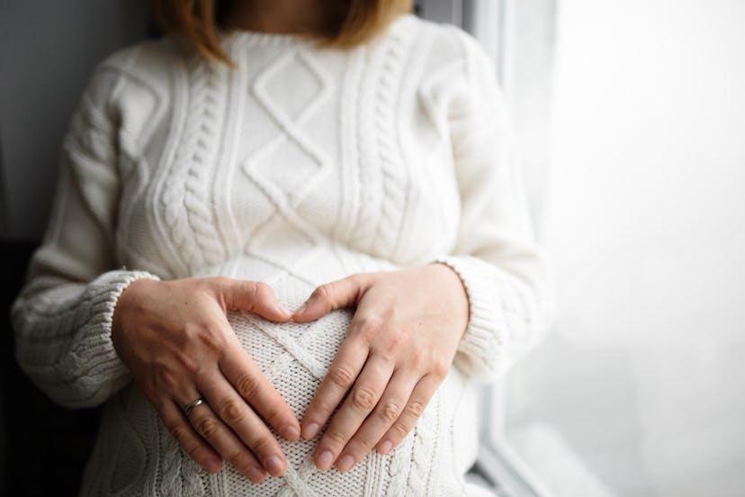 Hands in shape of heart on belly of pregnant woman in white warm knitted dress