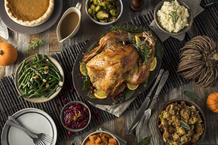 A Thanksgiving turkey is in the middle of a dinner table, surrounded by stuffing, cranberry sauce, a...