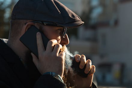 Vape bearded man in real life. Portrait of young guy with large beard in glasses and a gray cap talk...