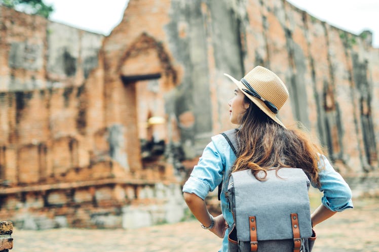 A woman wearing a blue backpack and tan straw hat with her back to the camera, explores ancient ruin...