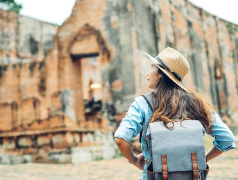 A woman wearing a blue backpack and tan straw hat with her back to the camera, explores ancient ruin...