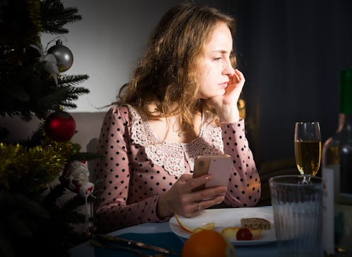 Upset woman sitting at table celebrating holiday alone and using phone at Christmas night