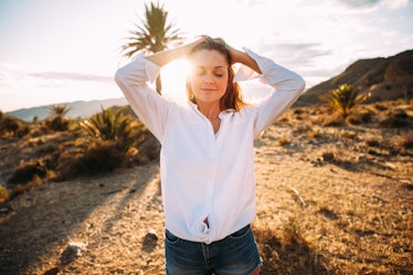 A woman in a white button-down smiles in a desert landscape with a palm tree and mountains in the ba...