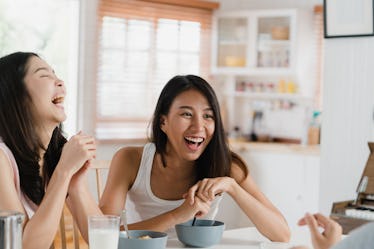 Two women laugh while eating breakfast in a bright kitchen at home.