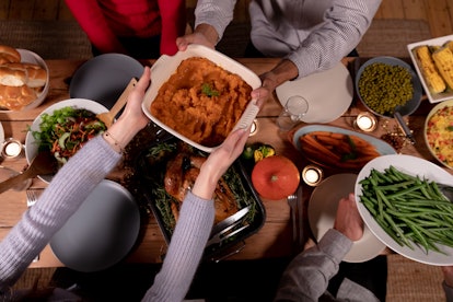 A group of friends sit around the dinner table and pass the side dishes to each other on Thanksgivin...