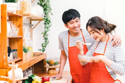 Young Asian lovely couple cooking together at home kitchen, wear red apron making lunch meal. Girl t...