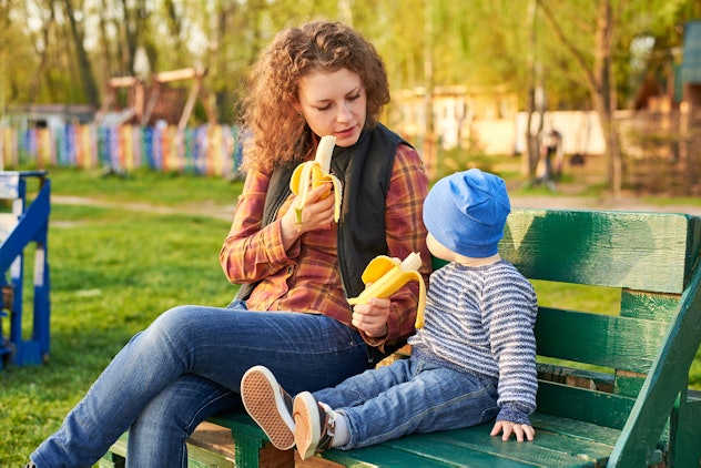 Mom eats and feeds her little son with banana on a bench in the park. Healthy eating concept