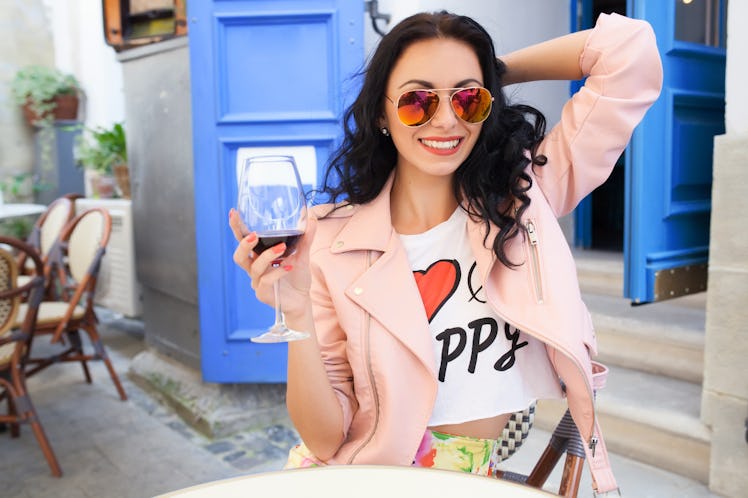 A happy woman with brown hair lifts her glass of red wine while relaxing at an outdoor restaurant.