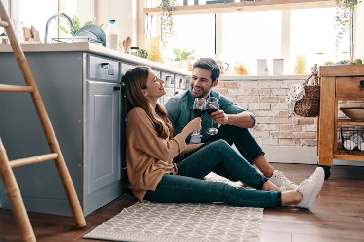 A happy couple toast their wine glasses on the kitchen floor.