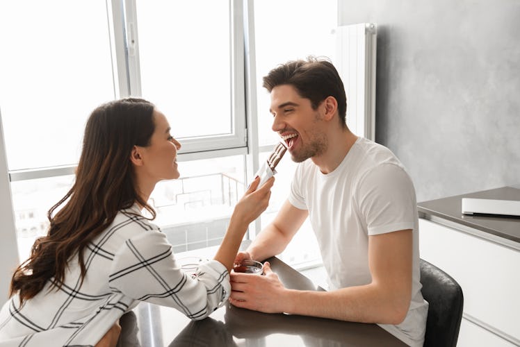 Young couple man and woman eating chocolate and drinking tea while sitting at table at home near big...