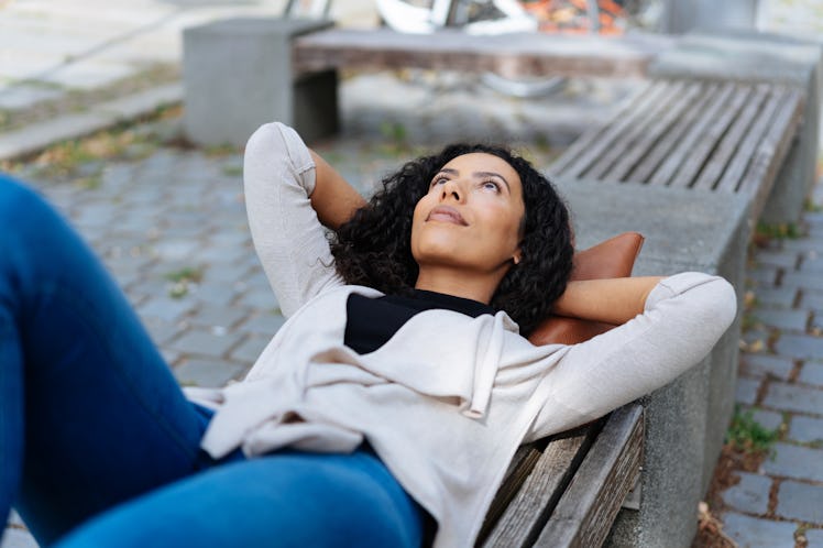 Young woman lying on a bench in an urban park or street daydreaming with her hands behind her head s...